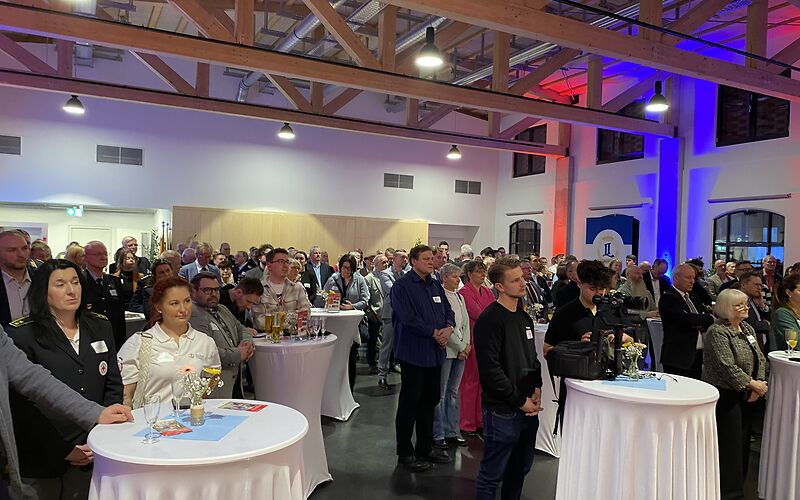 Blick von vorn in den gefüllten Saal, die Gäste stehen in Gruppen an Stehtischen mit weißen Tischdecken.