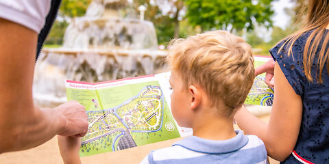 Mittig steht ein kleiner Junge, der auf einen Lageplan des Rosengartens schaut. Neben ihm stehen ein Mädchen und eine erwachsene Person, die jedoch nur teilweise zu sehen sind. Im Hintergrund ist der Kaskadenbrunnen.