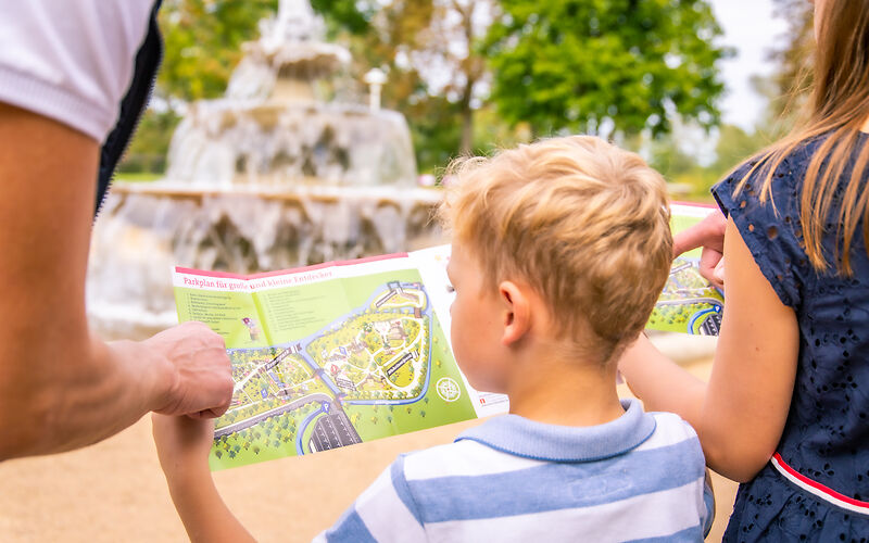 Mittig steht ein kleiner Junge, der auf einen Lageplan des Rosengartens schaut. Neben ihm stehen ein Mädchen und eine erwachsene Person, die jedoch nur teilweise zu sehen sind. Im Hintergrund ist der Kaskadenbrunnen.