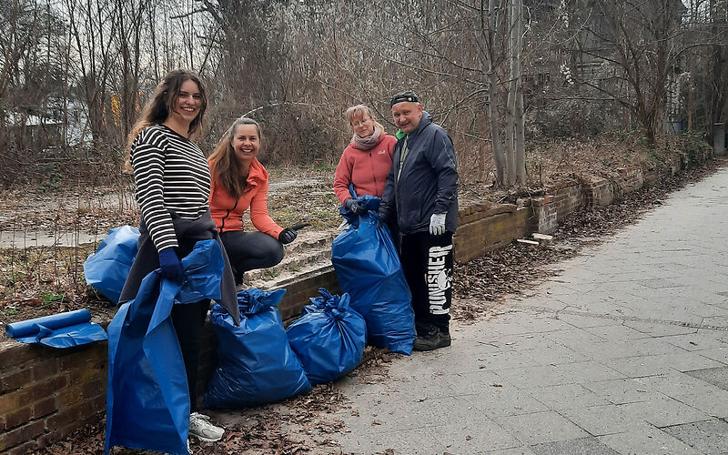 4 erwachsene Personen stehen mit mehreren  Müllsäcken vor einer kleinen Mauer neben einem geplasterten Weg. An der Mauer entlang liegt Laub.