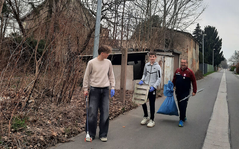 3 Personen gehen eine Staße entlang. Sie tragen eine Greifzange, einen blauen Müllsack und einen dreckigen Sitz eines Stuhles.