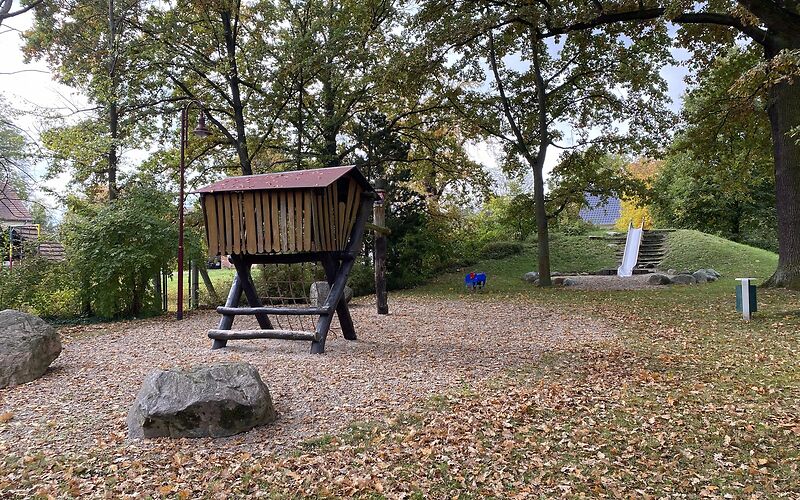 Auf einer Sandfläche steht ein Holzklettergerüst, der obere Teil ist ein Haus auf dicken Balken, dass über eine Kletternetz erreicht werden kann. Im Hintergrund steht am Rand der Fläche eine blaue Federwippe.