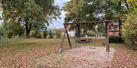 Zwei Schaukeln hängen nebeneinander an einem Holzgestell, welches sich auf einer Sandfläche befindet. 
