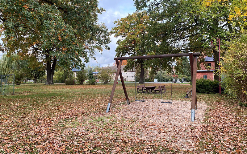 Zwei Schaukeln hängen nebeneinander an einem Holzgestell, welches sich auf einer Sandfläche befindet. 