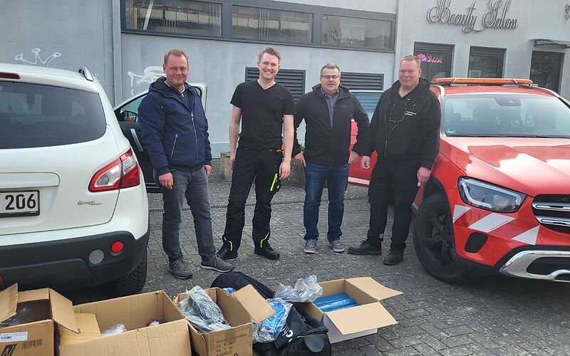 4 Männer in dunkler Kleidung auf einem Parkplatz. Vor Ihnen stehen Pappkisten mit Untensilien, z. B. blaue Müllsäcke.