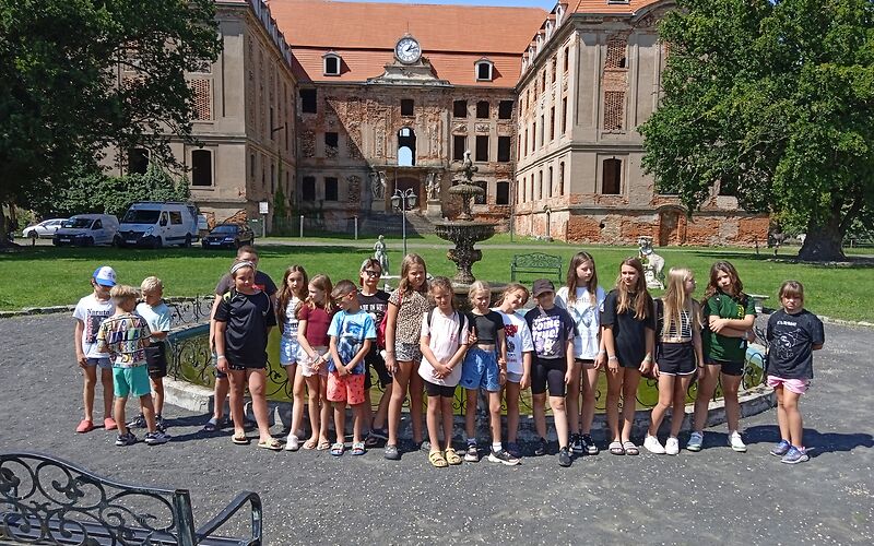 Die Schülerinnen und Schüler stehen in einer Reihe vor einem Brunnen. Im Hintergrund befindet sich die Schlossruine des Schlosses in Brody. Das Schloss ist ein U-Förmiges Gebäude mit einer großen Uhr in der Mitte. Es steht auf einer Wiese, links und rechts befinden sich große Bäume.
