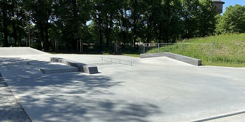 Leere Skateanlage mit Betonflächen, Rampen und Geländer, umgeben von Bäumen und einem Turm im Hintergrund