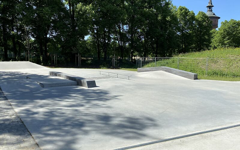 Leere Skateanlage mit Betonflächen, Rampen und Geländer, umgeben von Bäumen und einem Turm im Hintergrund