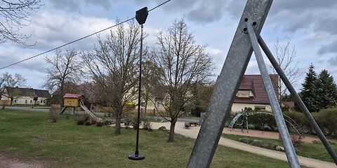 Im Vordergrund ist der Startpunkt der Seilbahn aus Metallstäben, an denen ein Seil befestigt ist. Der Sitz ist ebenfalls im Bild. Im Hintergrund sind sind ein Klettergerüst und eine Rutsche zu sehen. Der Spielplatz befindet sich auf einer Wiese und ist von Häusern umgeben.
