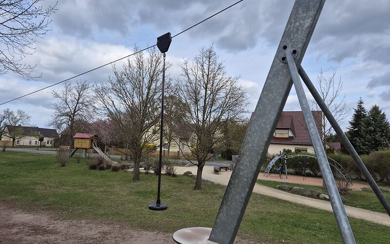 Im Vordergrund ist der Startpunkt der Seilbahn aus Metallstäben, an denen ein Seil befestigt ist. Der Sitz ist ebenfalls im Bild. Im Hintergrund sind sind ein Klettergerüst und eine Rutsche zu sehen. Der Spielplatz befindet sich auf einer Wiese und ist von Häusern umgeben.