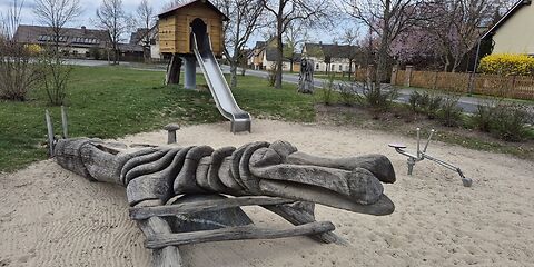 Im Sandbereich des Spielplatzes stehen ein Bagger und eine hölzerne längliche Figur zum Klettern. Es kann nicht genau definiert werden, was sie darstellt. Dahinter ist eine Holzhütte auf einer Runden Säule. Aus der Hütte hinaus führt eine Rutsche. Im Hintergrund befinden sich Wiese, eine Straße und Häuser.