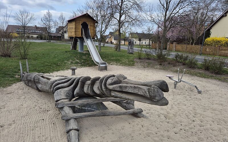 Im Sandbereich des Spielplatzes stehen ein Bagger und eine hölzerne längliche Figur zum Klettern. Es kann nicht genau definiert werden, was sie darstellt. Dahinter ist eine Holzhütte auf einer Runden Säule. Aus der Hütte hinaus führt eine Rutsche. Im Hintergrund befinden sich Wiese, eine Straße und Häuser.