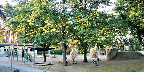 Zu sehen ist der Spielplatz der Kita, Kletterfelsen und hoher Baumbestand, Wippen, ein Sandkasten und eine Rutsche