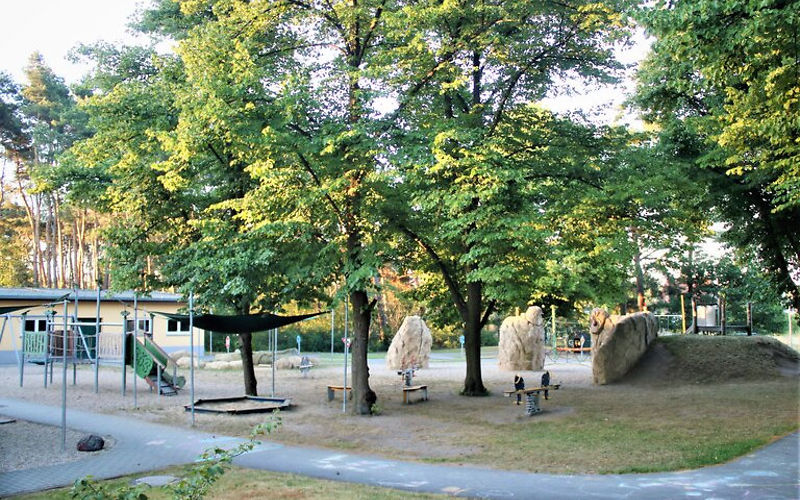Zu sehen ist der Spielplatz der Kita, Kletterfelsen und hoher Baumbestand, Wippen, ein Sandkasten und eine Rutsche