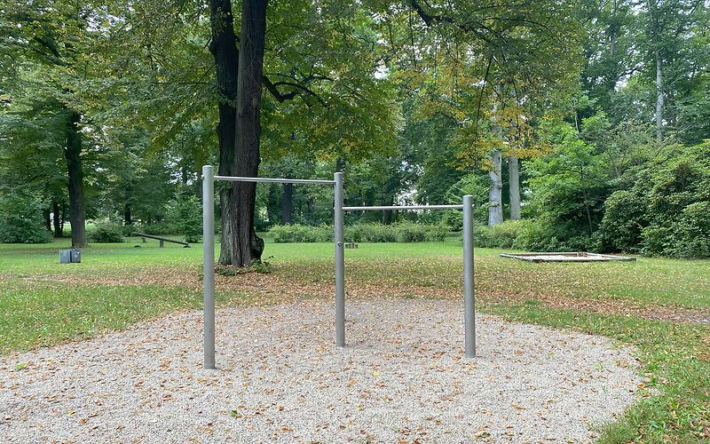 Spielgerät aus 3 Pfeilern sind oben auf 2 unterschiedlichen Höhen Metallstangen befestigt. Das Spielgerät steht auf einer Sandfläche.