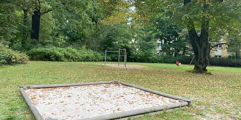 Ein Sandkasten mit Holzumrandung auf einer Wiese. Im Sand und auf der Wiese liegen bunte Blätter.