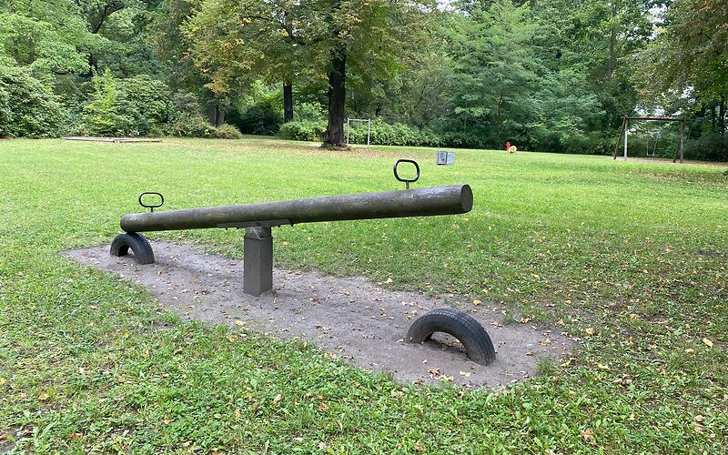 Eine Holzwippe auf einer Sandfläche. An jeder Seite der Wippe ist ein Griff und unter der Wippe ein zur Hälfte eingegrabener Reifen.