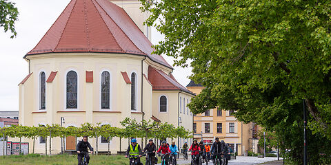 Die Gruppe fährt mit Fahrrädern auf einer Straße, im Hintergrund links sieht man den Platanenhain und die Stadtkirche.