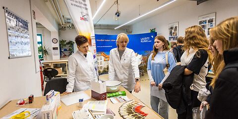 Gruppe von Jugendlichen Informiert sich am Stand der Rosenapotheke, an dem Infomaterialien auf einem großen Holztisch liegen, hinter dem 2 Mitarbeiterinnen im Apotheker-Kittel stehen.