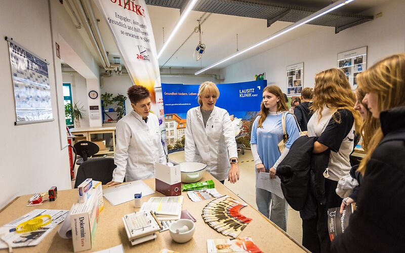 Gruppe von Jugendlichen Informiert sich am Stand der Rosenapotheke, an dem Infomaterialien auf einem großen Holztisch liegen, hinter dem 2 Mitarbeiterinnen im Apotheker-Kittel stehen.