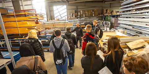 Gruppen von Jugendlichen besichtigt eine Lagerhalle, in der Holz- und Metall aufbewahrt wird, im Hintergrund stehen Kisten auf einem Regal. Die Halle hat zwei große Fenster.