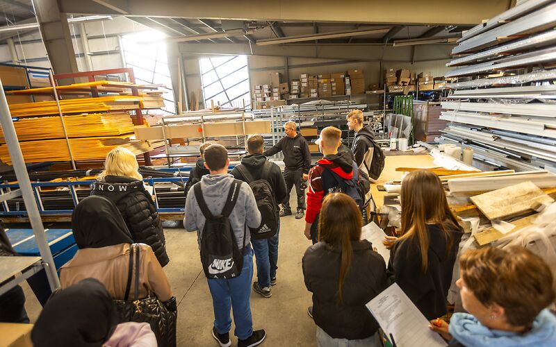 Gruppen von Jugendlichen besichtigt eine Lagerhalle, in der Holz- und Metall aufbewahrt wird, im Hintergrund stehen Kisten auf einem Regal. Die Halle hat zwei große Fenster.