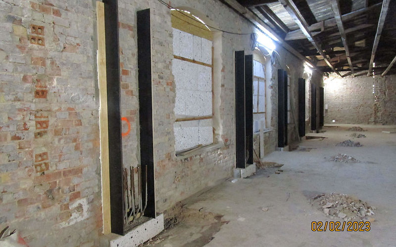 Zu sehen sind mit Holzbrettern eingeschalte Wandvorlagen, welche noch betoniert werden. Zwischen den Wandvorlagen sind abgedeckte Industriefenster zu erkennen. Am noch nicht sanierten Boden liegen Baumaterialien.