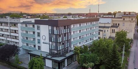 Zu sehen ist das Gebäude Quartier am Berliner Platz, ein Eckgebäude, im Bereich des Erdgeschosses sind große Schaufenster zu sehen, darüberliegend 4 Etagen Wohnungen mit Fenstern und Balkonen