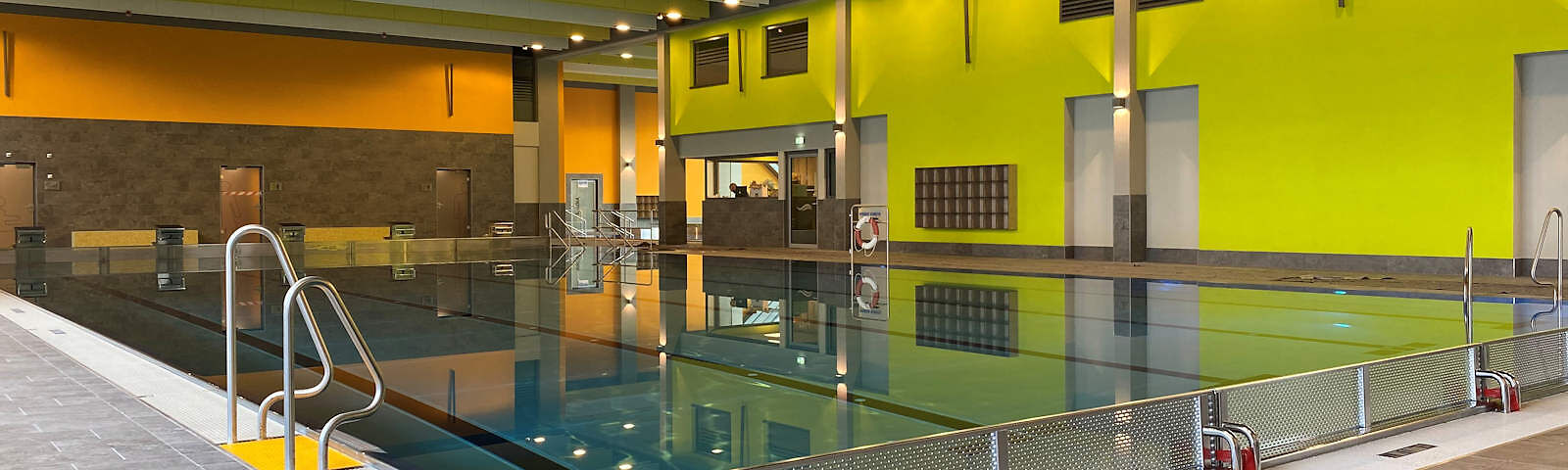 Blick auf ein 5-bahniges Schwimmbecken mit Ein- und Austeigen an den Seiten (hinten eine Treppe und zwei Leitern). Die Wand rechts ist grün gestrichen, die Wand links ist unten dunkelbraun und oben orange.