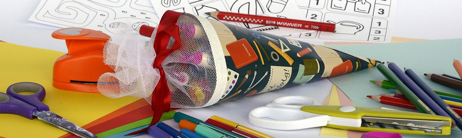 Eine Schulzuckertüte liegt auf einem Tisch umgeben von Stiftenm Papier, einer Schere, einem Lochstempel und Ausmalbildern