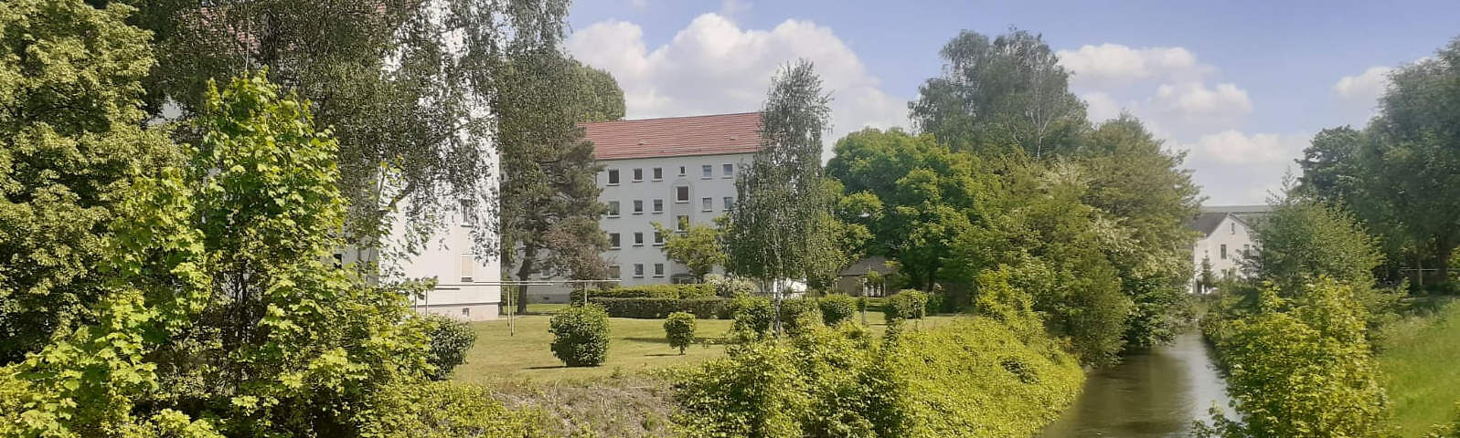Zwischen grünen Bäumen und Sträuchern stehen zwei weiße Häuser mit spitzem roten Dach, rechts im Bild ist ein schmaler Fluss.