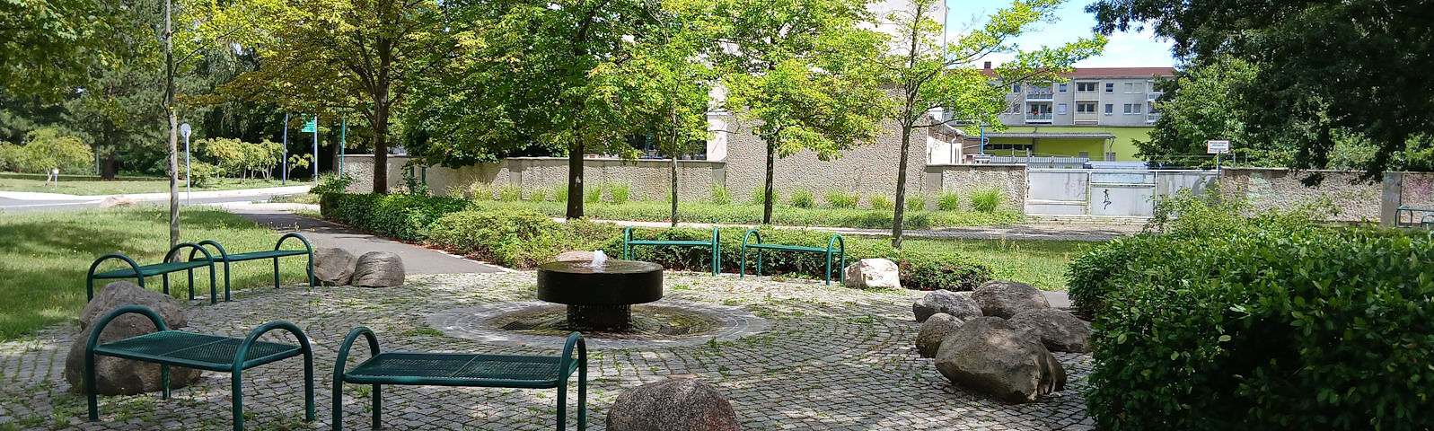 Sitzbereich mit im Kreis angeordneten Bänken um einen steinernden Springbrunnen herum in einer kleinen Parkanlage