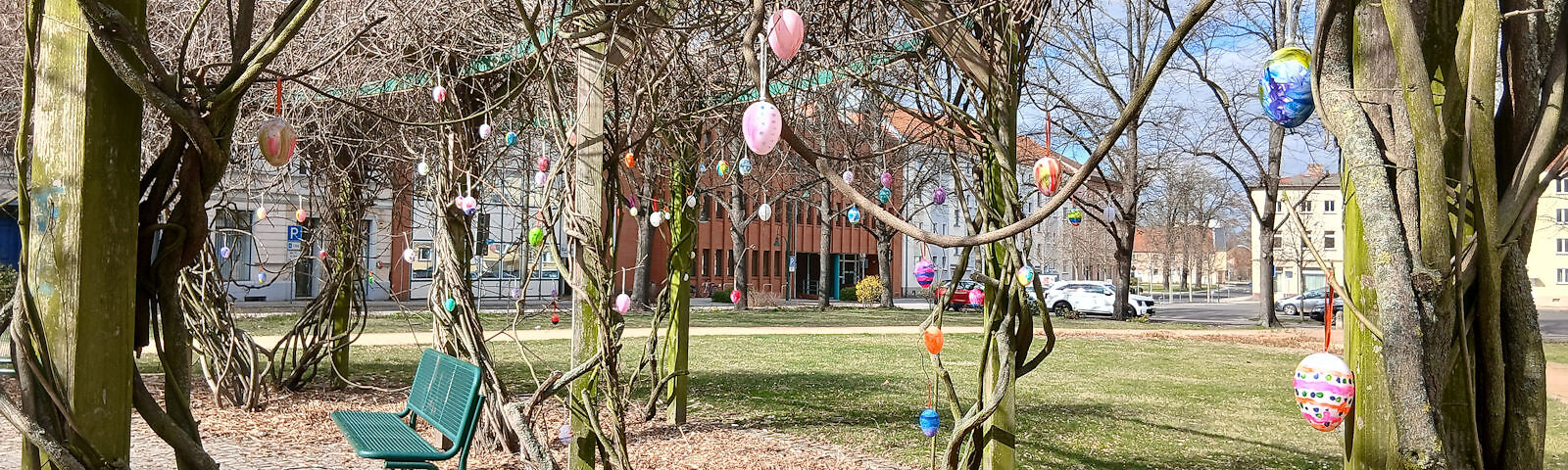 Park mit mehreren Bänken und Bäumen, an denen bunte Ostereier hängen, im Hintergrund das rote Rathausgebäude und die Stadtbibliothek 