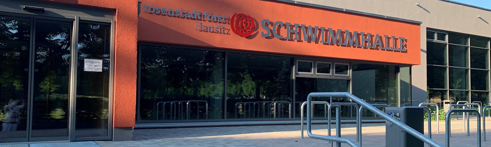 Eingangsbereich einer Schwimmhalle mit oranger Fassade, großen Fenstern und Schriftzug 'rosenstadt forst lausitz - SCHWIMMHALLE'