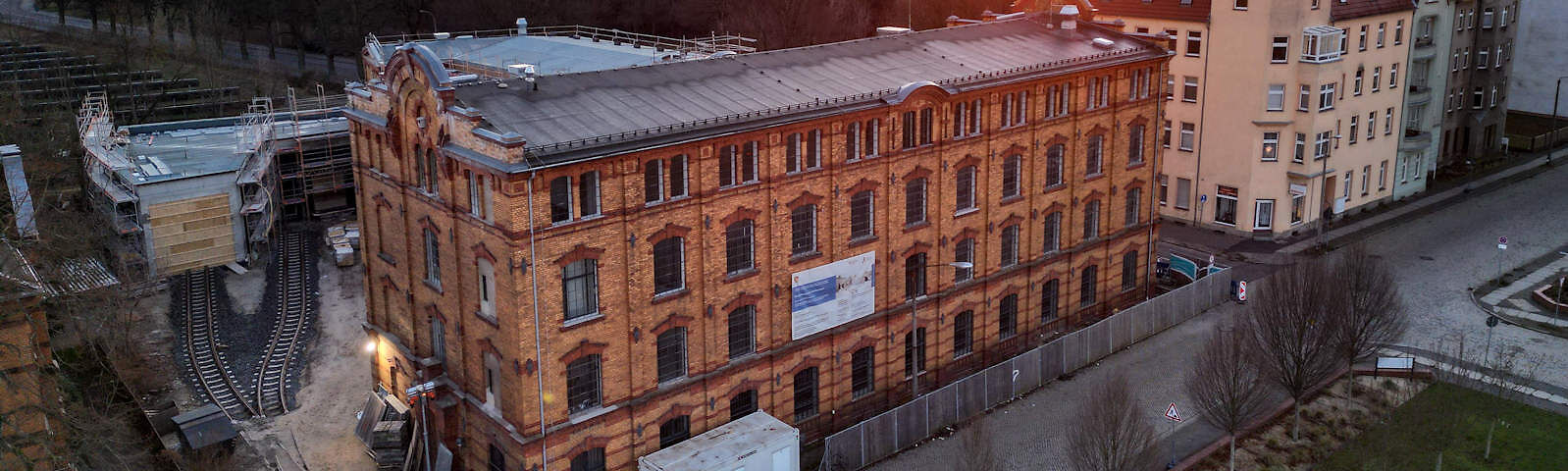 Vor dem Textilmuseum, einem alten Steingebäude, ist vor dem Gehweg ein Bauzaun aufgestellt worden. Das kleinere Nebengebäude hinter dem Museum ist mit einem Gerüst umstellt. Über den Hof des Museums verlaufen zwei Schienen, die in der Mitte des Hofes ineinander übergehen
